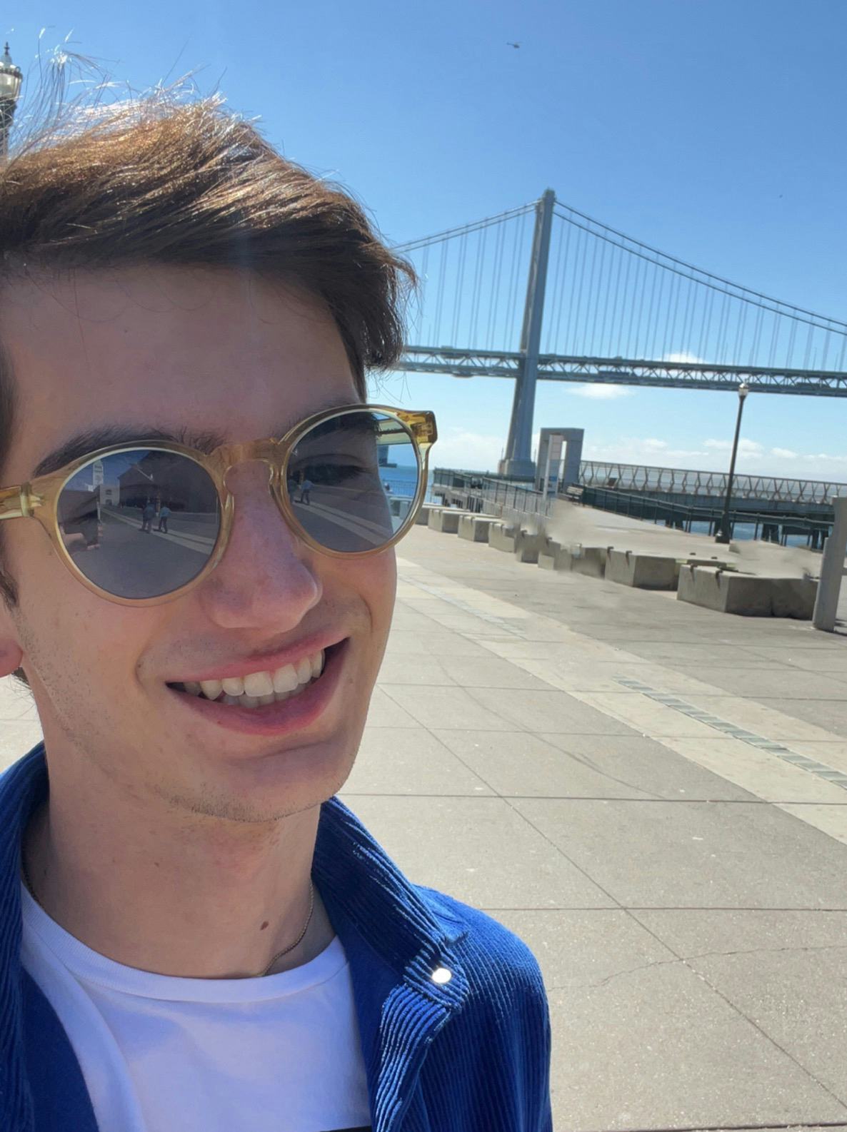 Me smiling wearing a deep blue curduroy shirt and yellow oversized round sunglasses in front of the Bay Bridge.