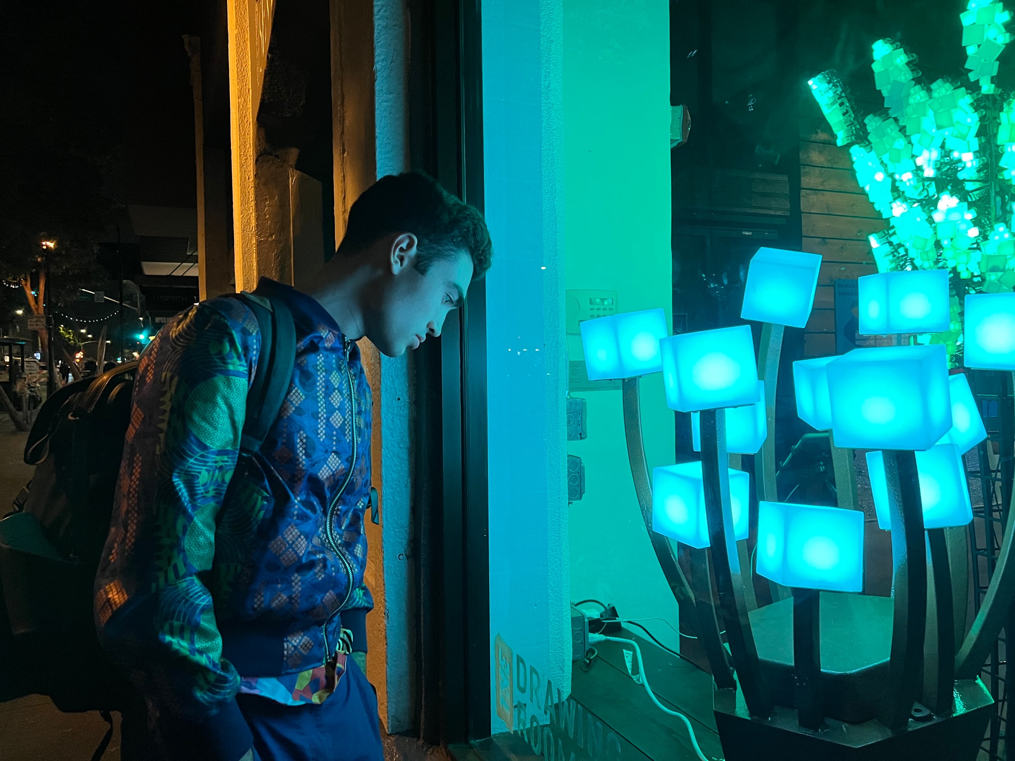 Lachlan wearing a reflective bomber jacket appearing moody looking at an art exhibit in a window with green lighting